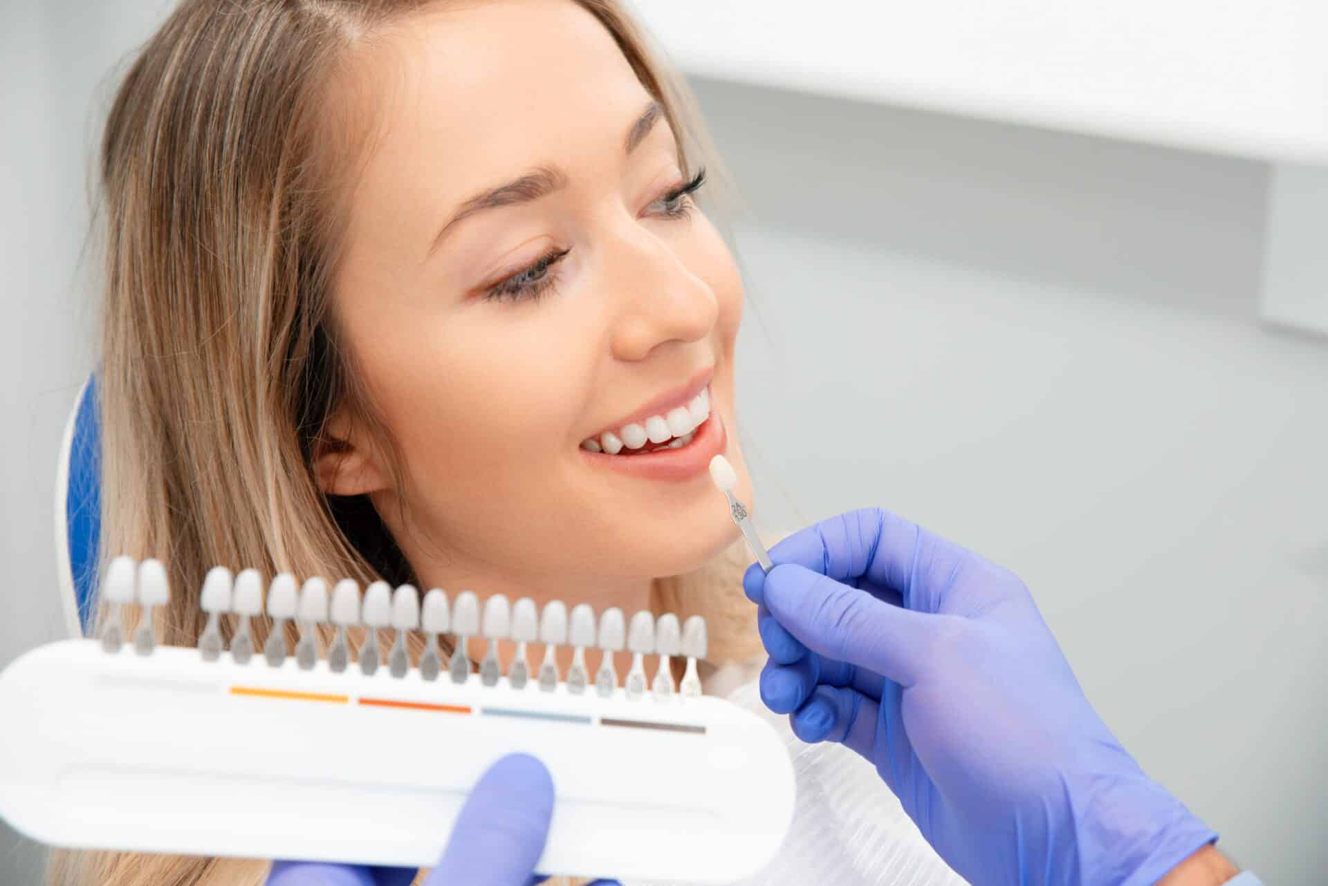 Dentist holds up shading palette of veneer samples to compare to woman's teeth while she sits and smiles in a dental chair.