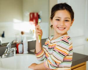 Young girl with brown hair and brown eyes wearing a colorful striped shirt holds up a toothbrush while smiling for the camera near her bathroom sink.