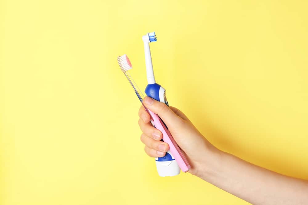 Hand holding an electric and manual toothbrush over a yellow background