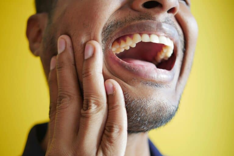 Close-up of man against yellow background holding the side of his face from dental pain due to a cracked crown.